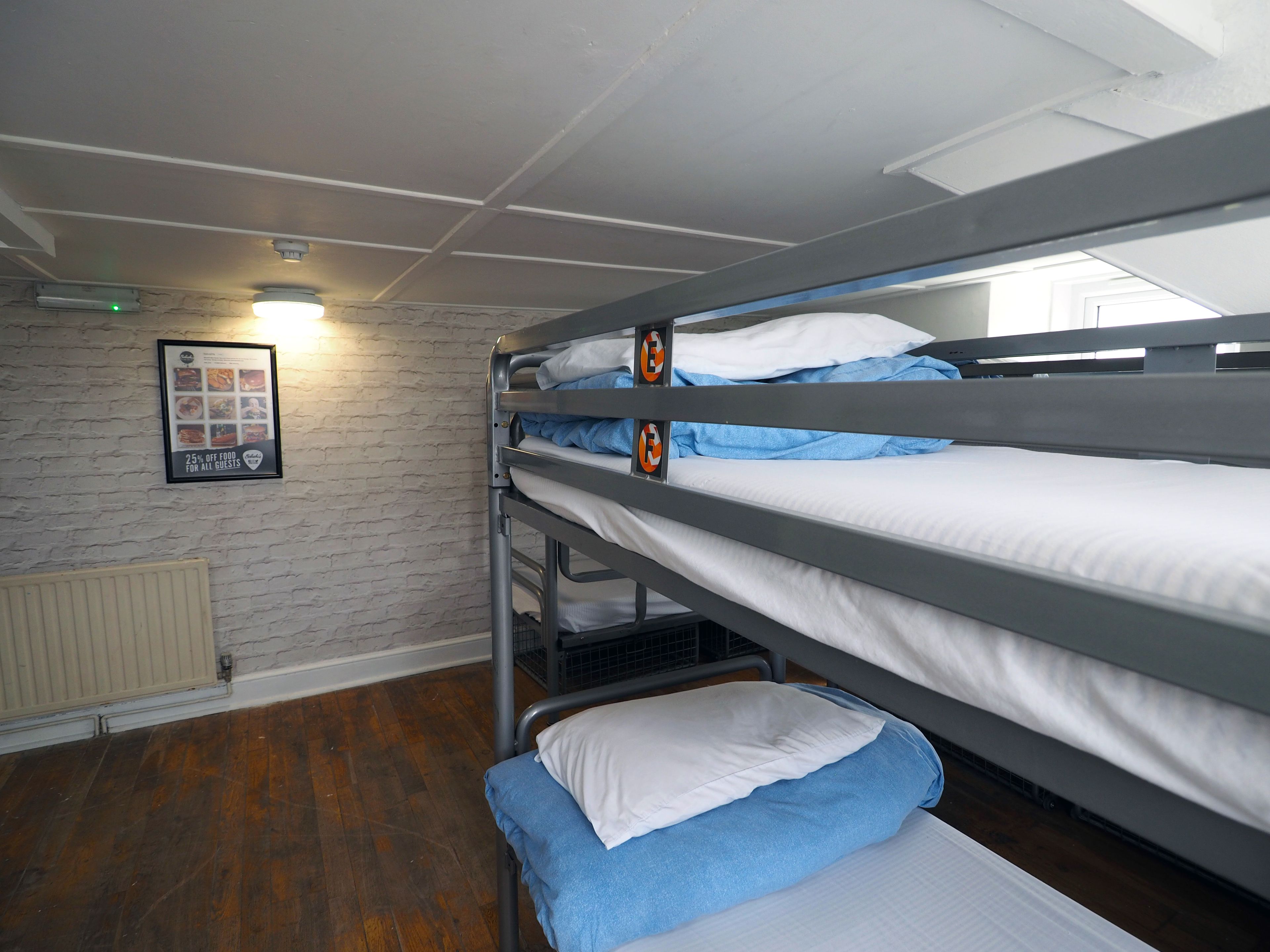 Une chambre avec des lits superposés, un menu encadré accroché au mur, un radiateur et un plancher en bois.