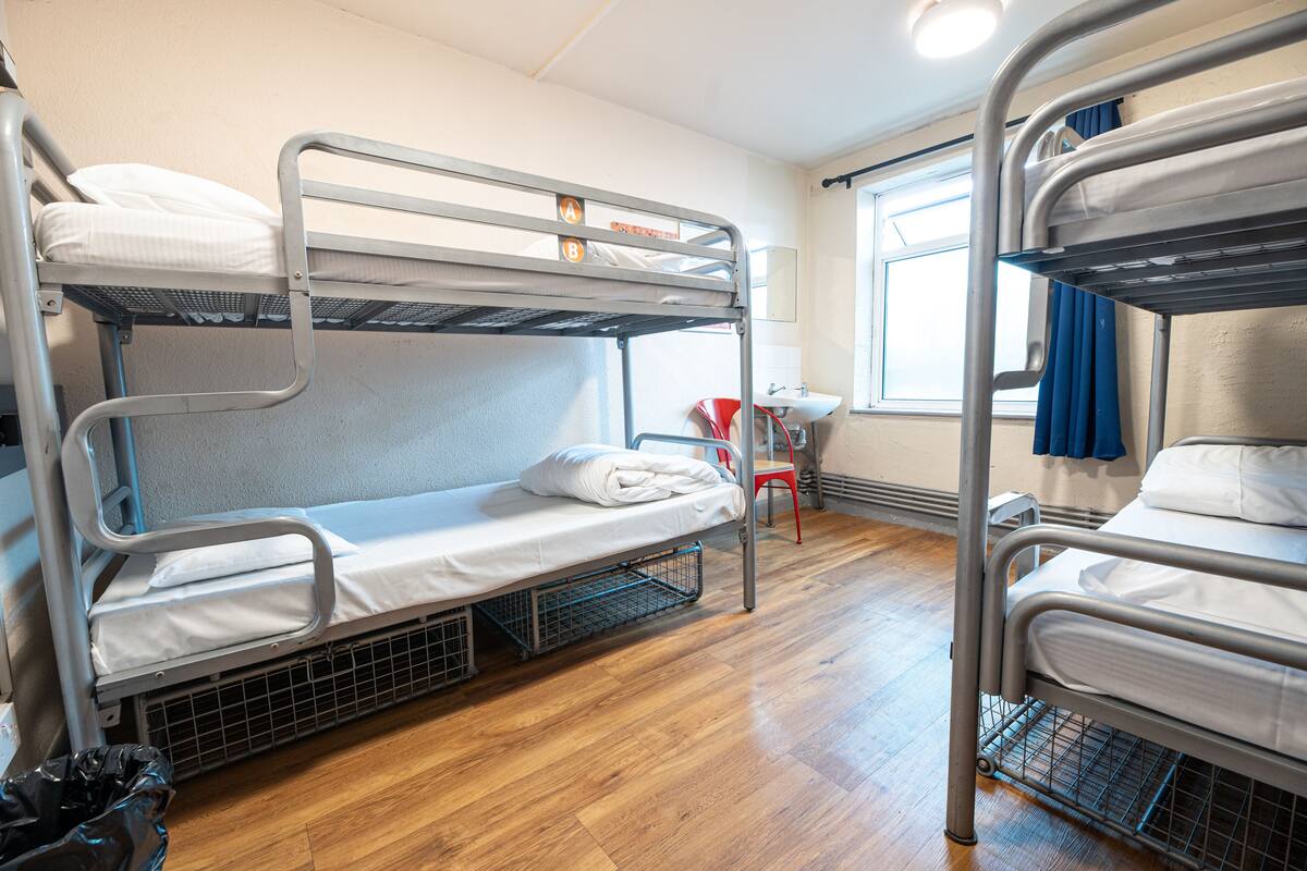 A bunk bed room with a small table and chair, a window with curtains, and a wooden floor.