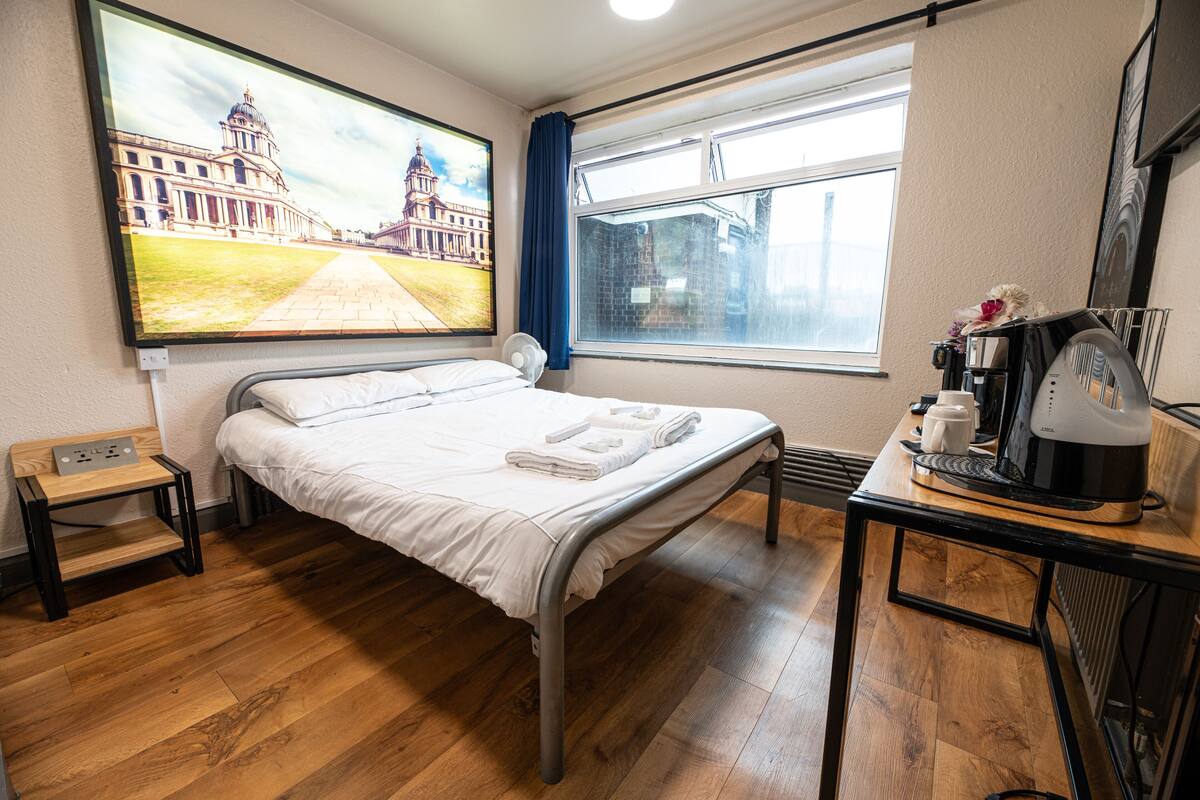 A hotel room with a bed, a small table with a coffee maker, a window with a view of a building, and a painting on the wall.