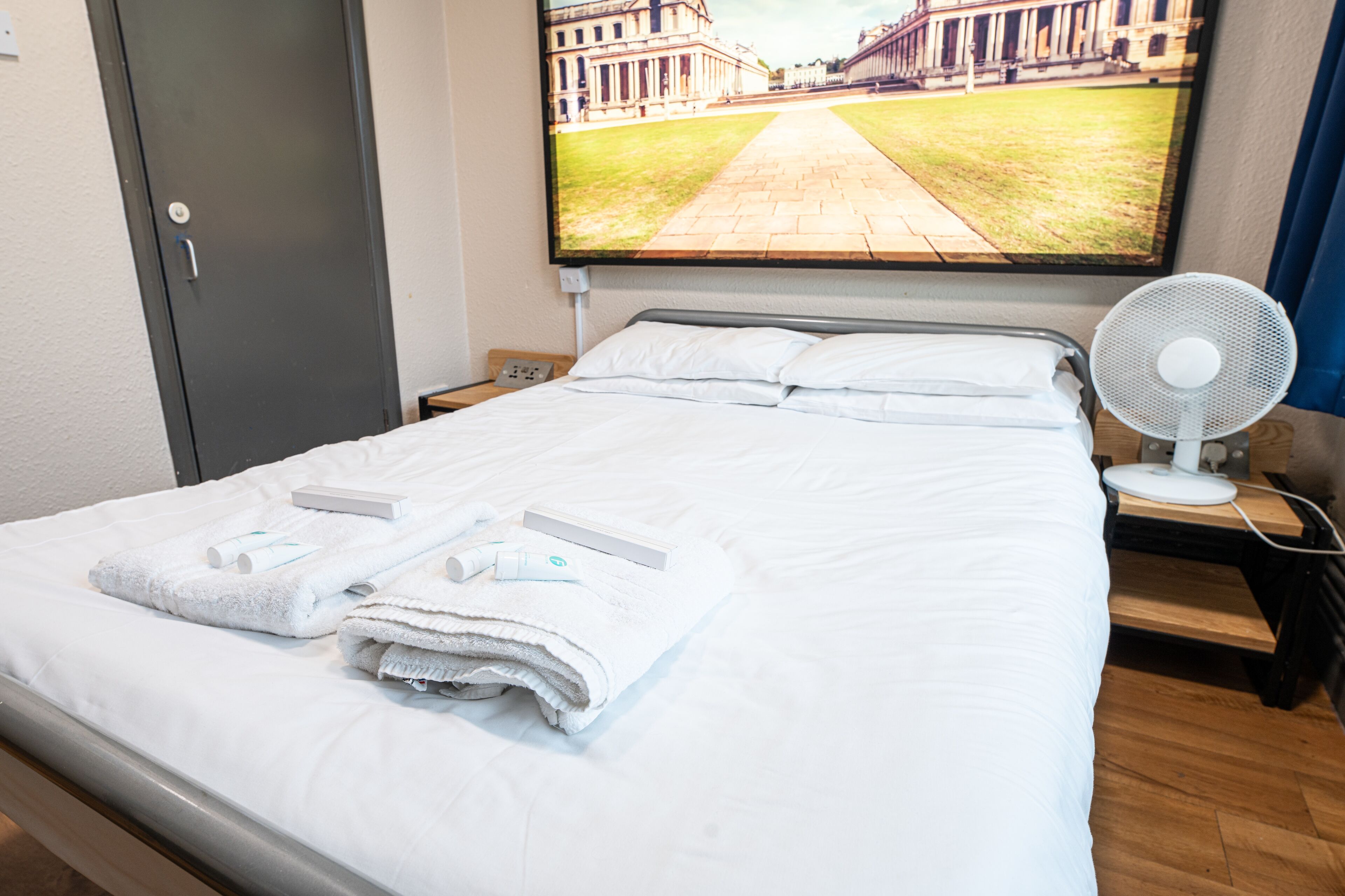 A hotel room with a neatly made bed, a bedside table with a fan, and a large window displaying a building view.
