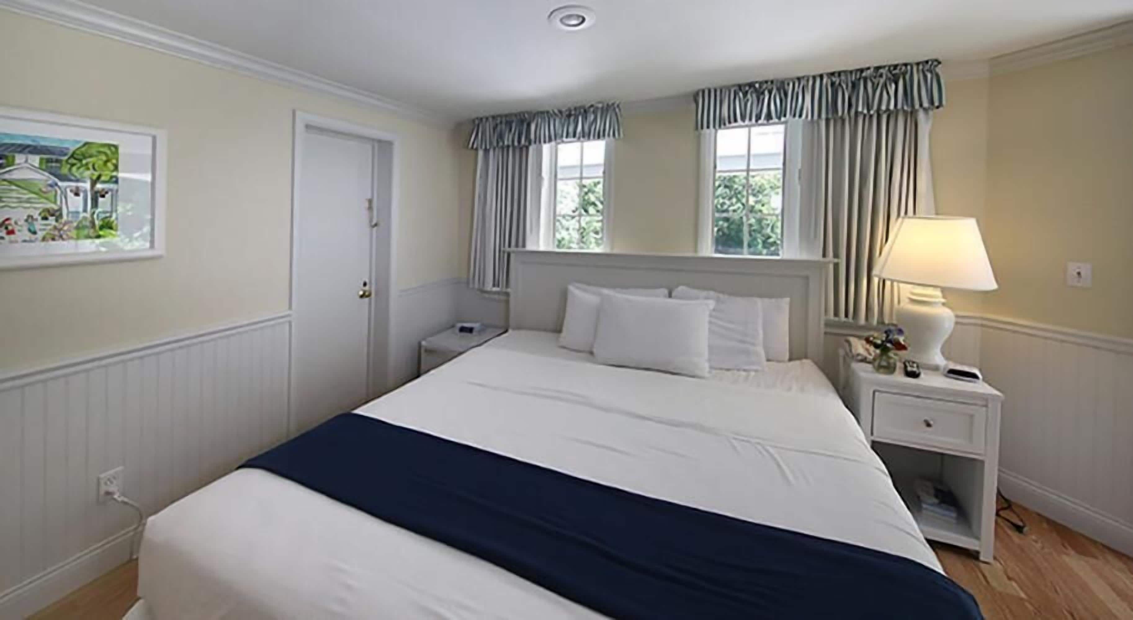 Une chambre d’hôtel bien rangée, avec un grand lit, une table de chevet avec une lampe et une fenêtre avec des rideaux.