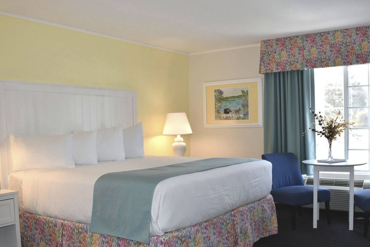 A hotel room with a bed, a desk, a chair, a lamp, a window with curtains, and a framed picture on the wall.