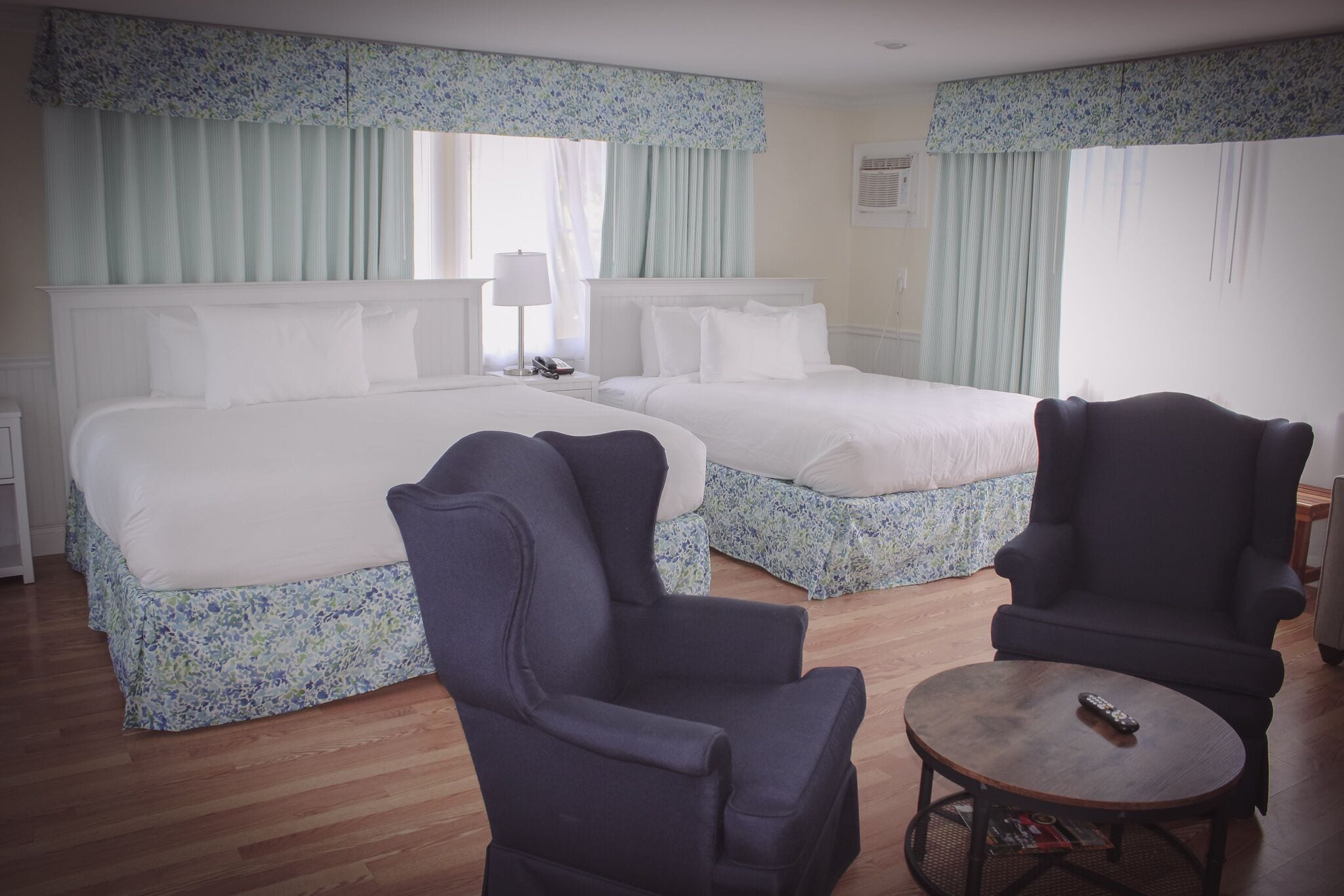 Habitación de hotel con dos camas, una mesa redonda, dos sillones y un ventanal con cortinas.