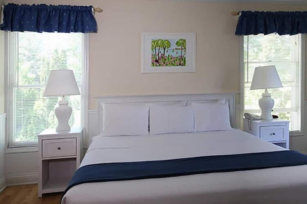 A bedroom with a large bed, two bedside tables with lamps, a framed picture on the wall, and windows with curtains.