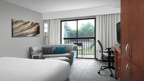 Habitación de hotel con una cama grande, un sofá, un escritorio con una silla, un televisor y un balcón con vistas a zonas verdes.