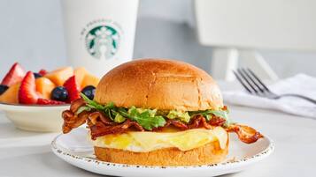 Un panino per la colazione con bacon, lattuga e formaggio su un panino, accompagnato da una ciotola di frutta e una tazza Starbucks.