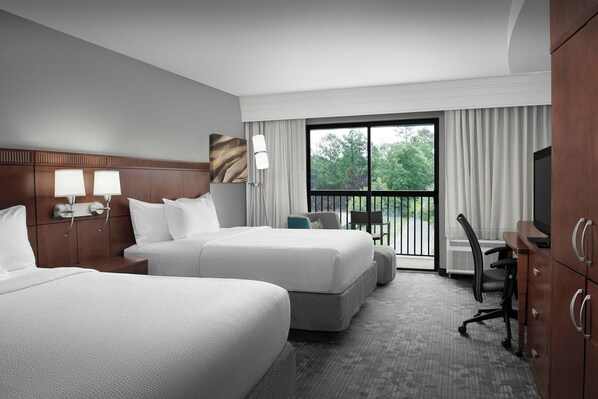 A hotel room with two beds, a desk, a chair, and a view of the outdoors.