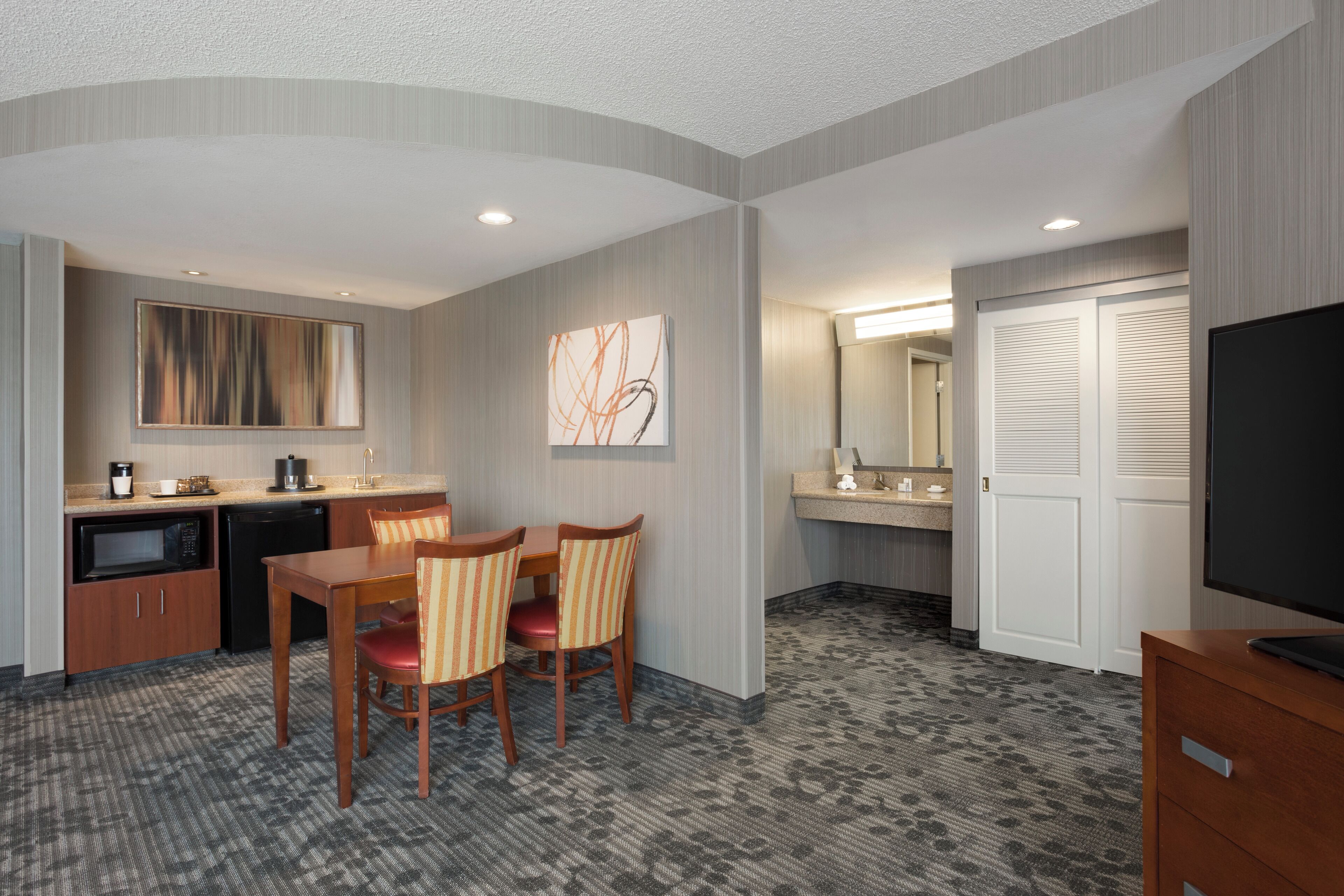A hotel room with a kitchenette, a dining area with chairs, a television, and a bathroom with a mirror and toilet.
