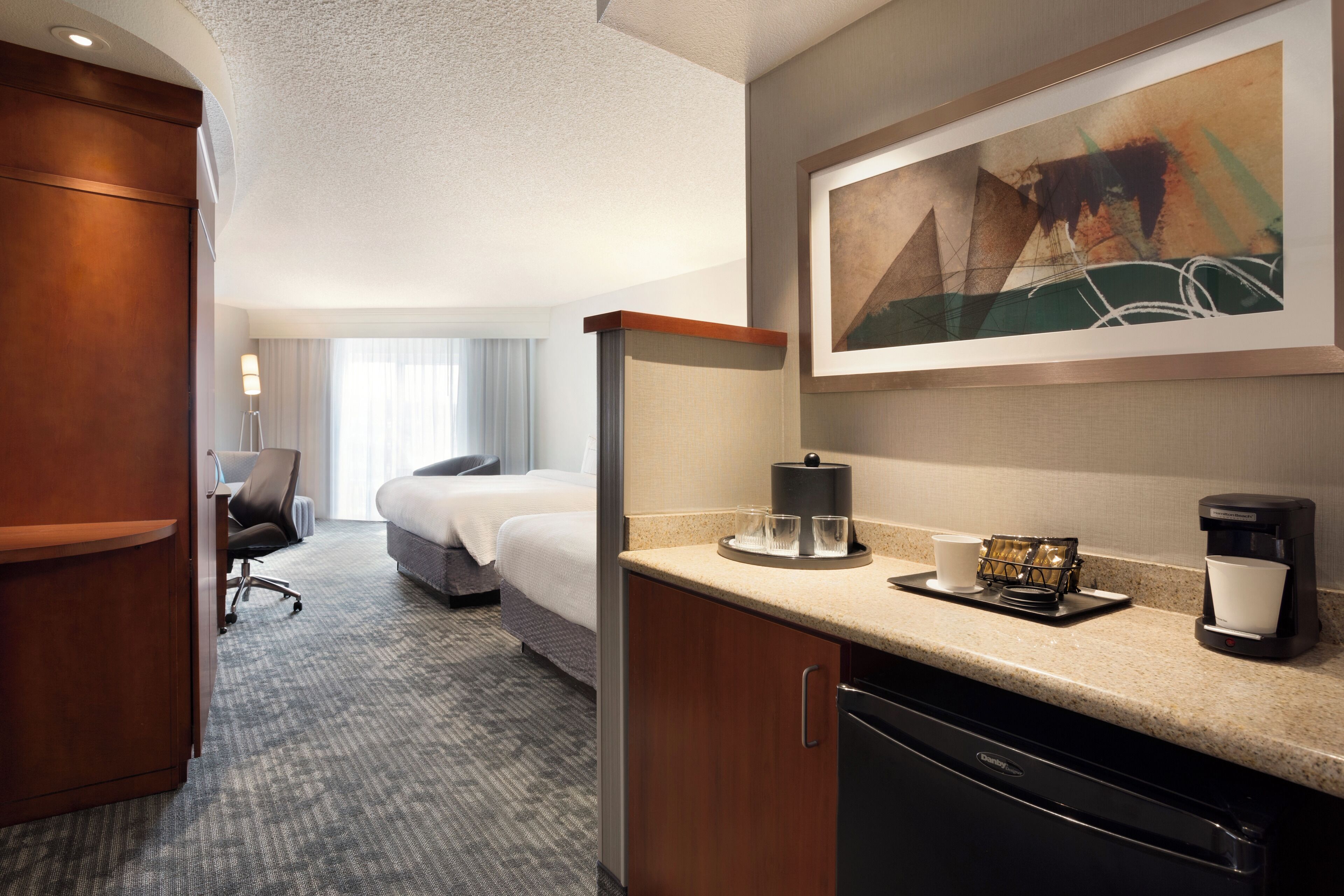 A hotel room with a kitchenette, a bed, a desk, and a large painting on the wall.