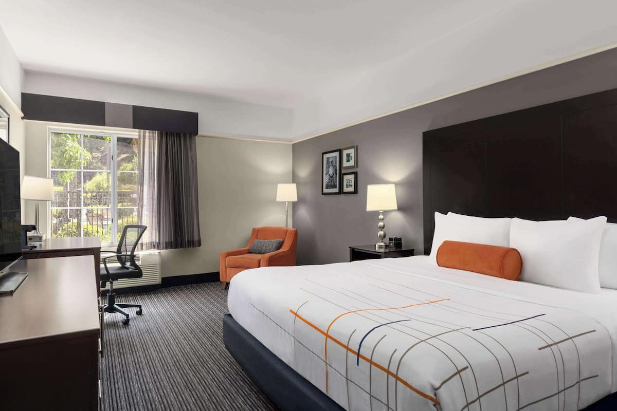 Ein Hotelzimmer mit einem großen Bett, einem Schreibtisch, einem Sessel, einem Fernseher und einem Fenster mit Vorhängen.