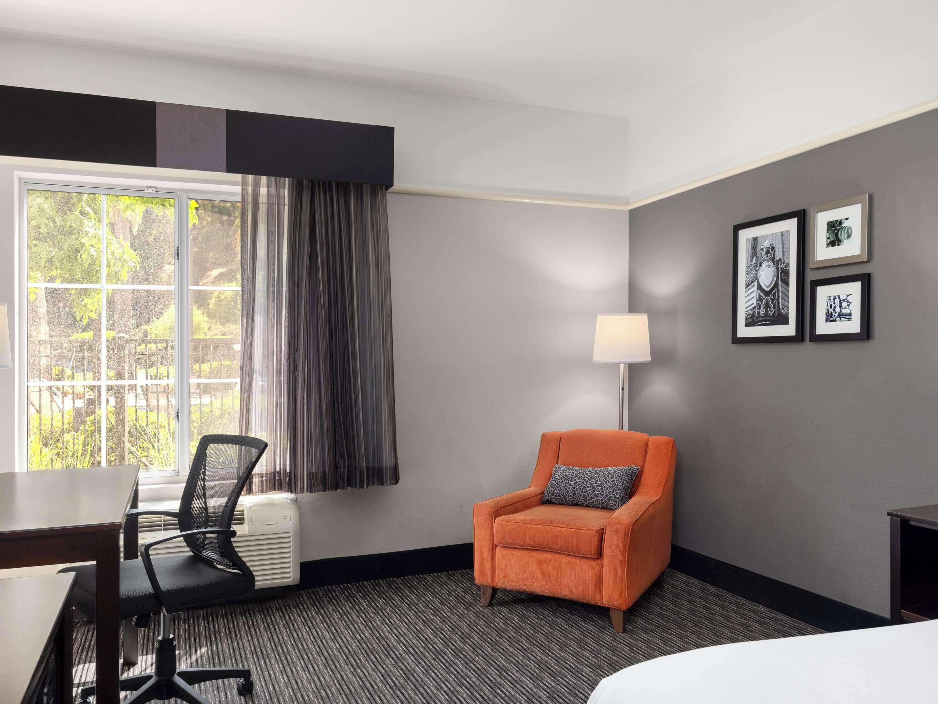 Una habitación de hotel con escritorio, silla y un sillón naranja. Hay una ventana con cortinas, una lámpara y cuadros en la pared.