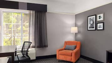 A hotel room with a desk, chair, and an orange armchair. There is a window with curtains, a lamp, and framed pictures on the wall.