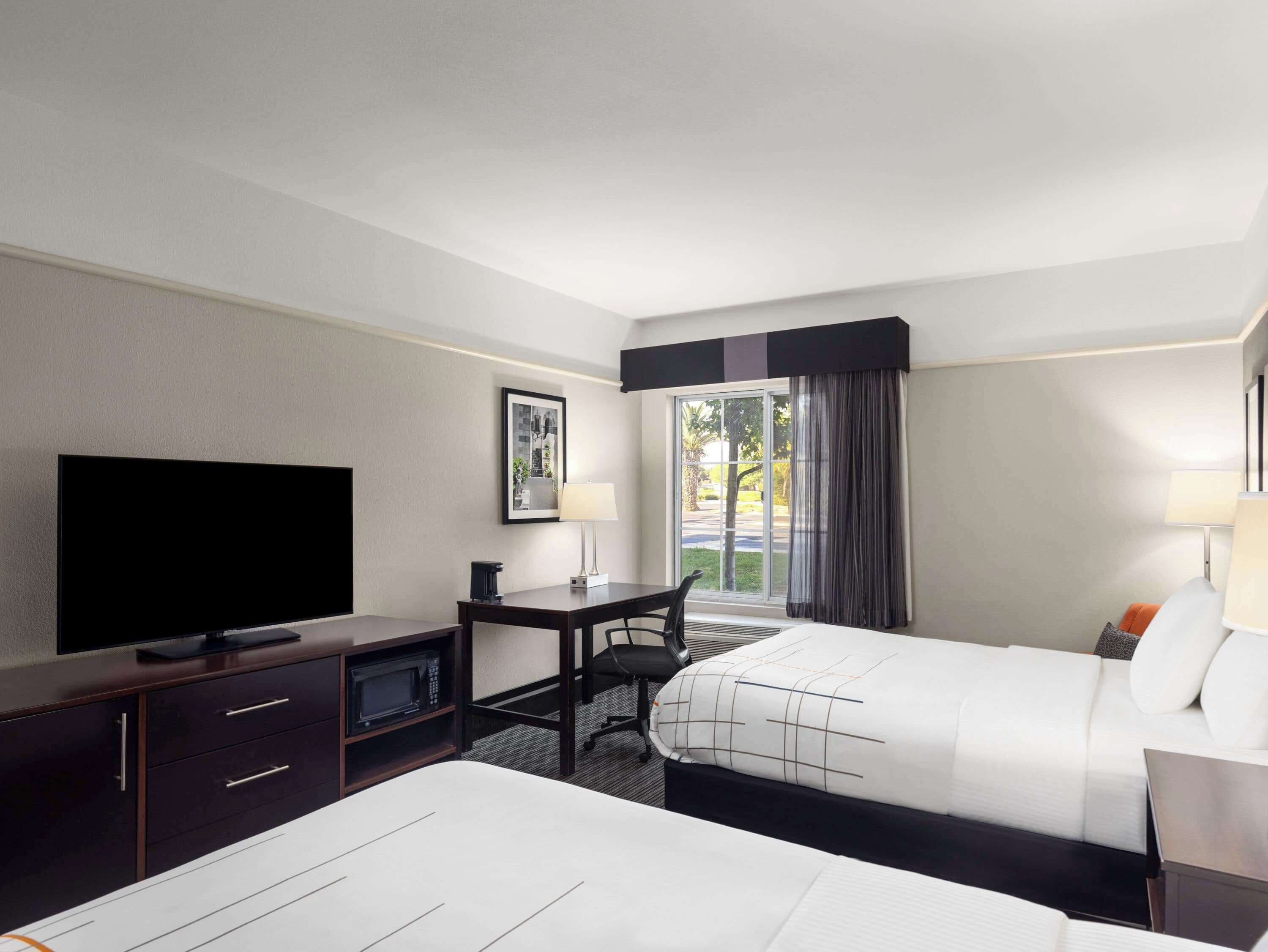 Une chambre d’hôtel avec un lit, un bureau avec un ordinateur, une télévision et une fenêtre avec des rideaux.
