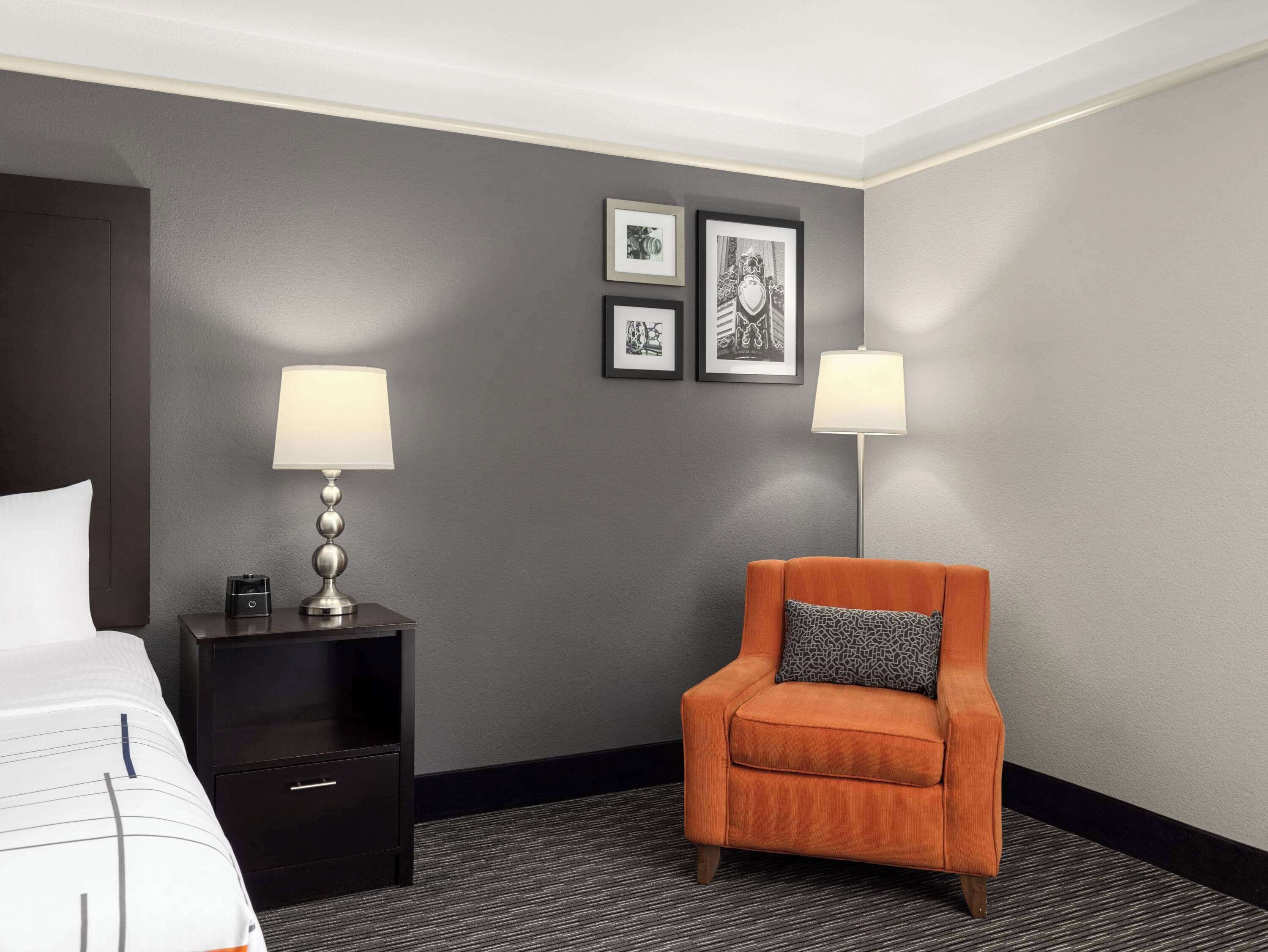 A hotel room with a bed, a nightstand with a lamp, an orange armchair, and framed pictures on the wall.