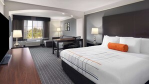 Ein modernes Hotelzimmer mit einem großen Bett, einem Schreibtisch, einem Stuhl und einem Sofa. Auf dem Schreibtisch stehen zwei Lampen und ein Fenster mit Vorhängen.