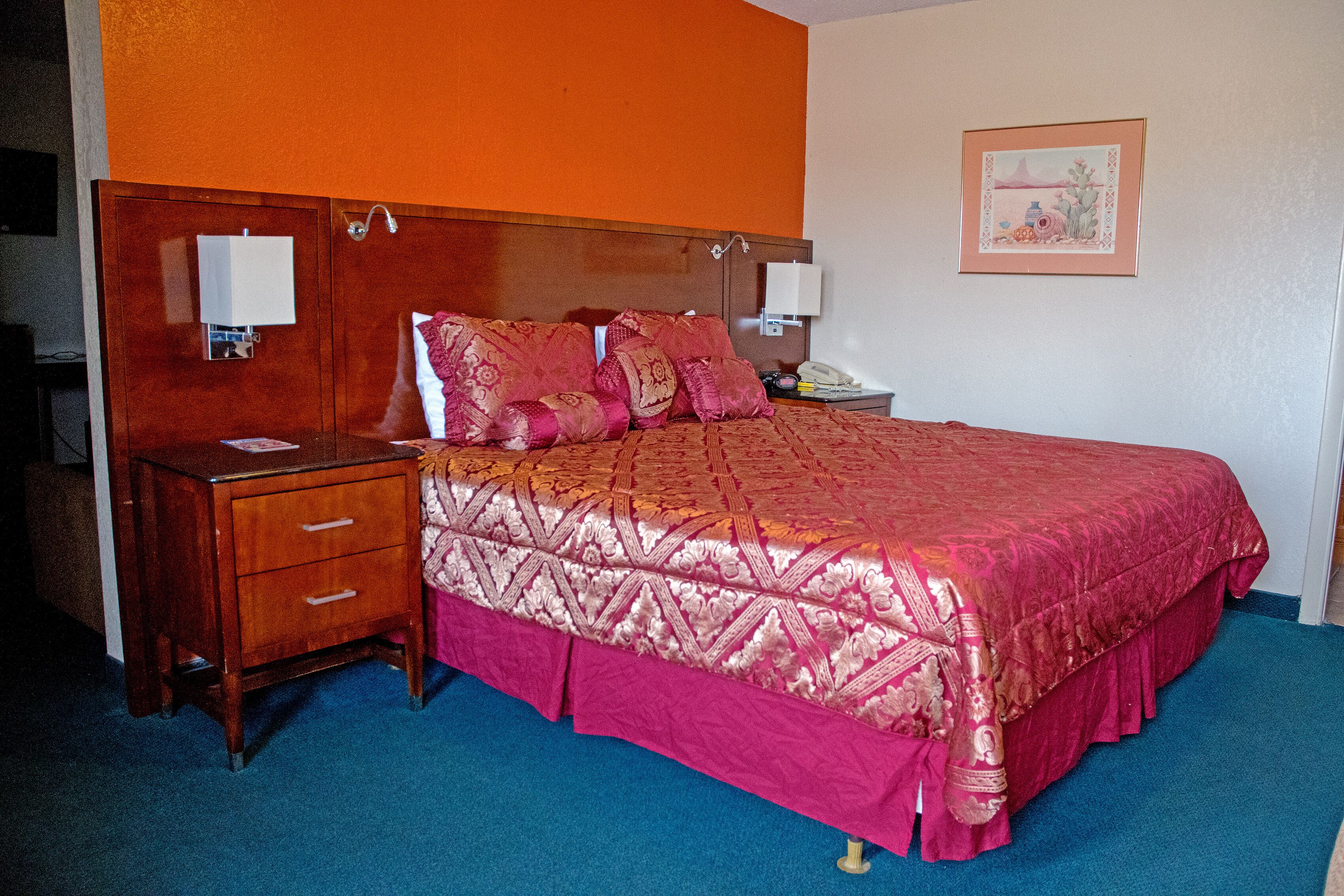 A hotel room with a wooden headboard, a bed with a pink and gold patterned bedspread, a nightstand with a lamp, and a framed picture on the wall.