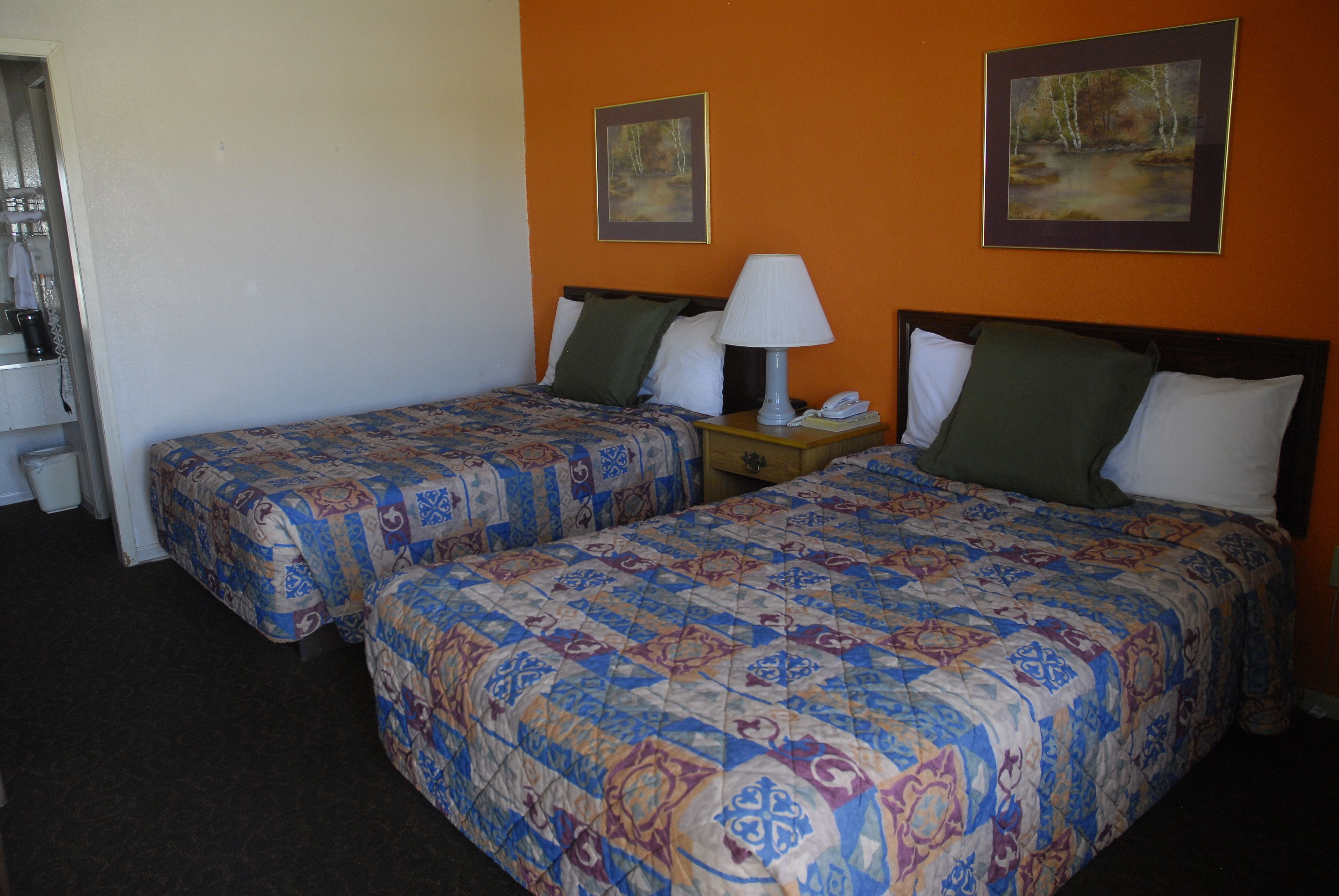 A hotel room with two beds, each with a patterned bedspread, a bedside table with a lamp, and a framed picture on the wall.