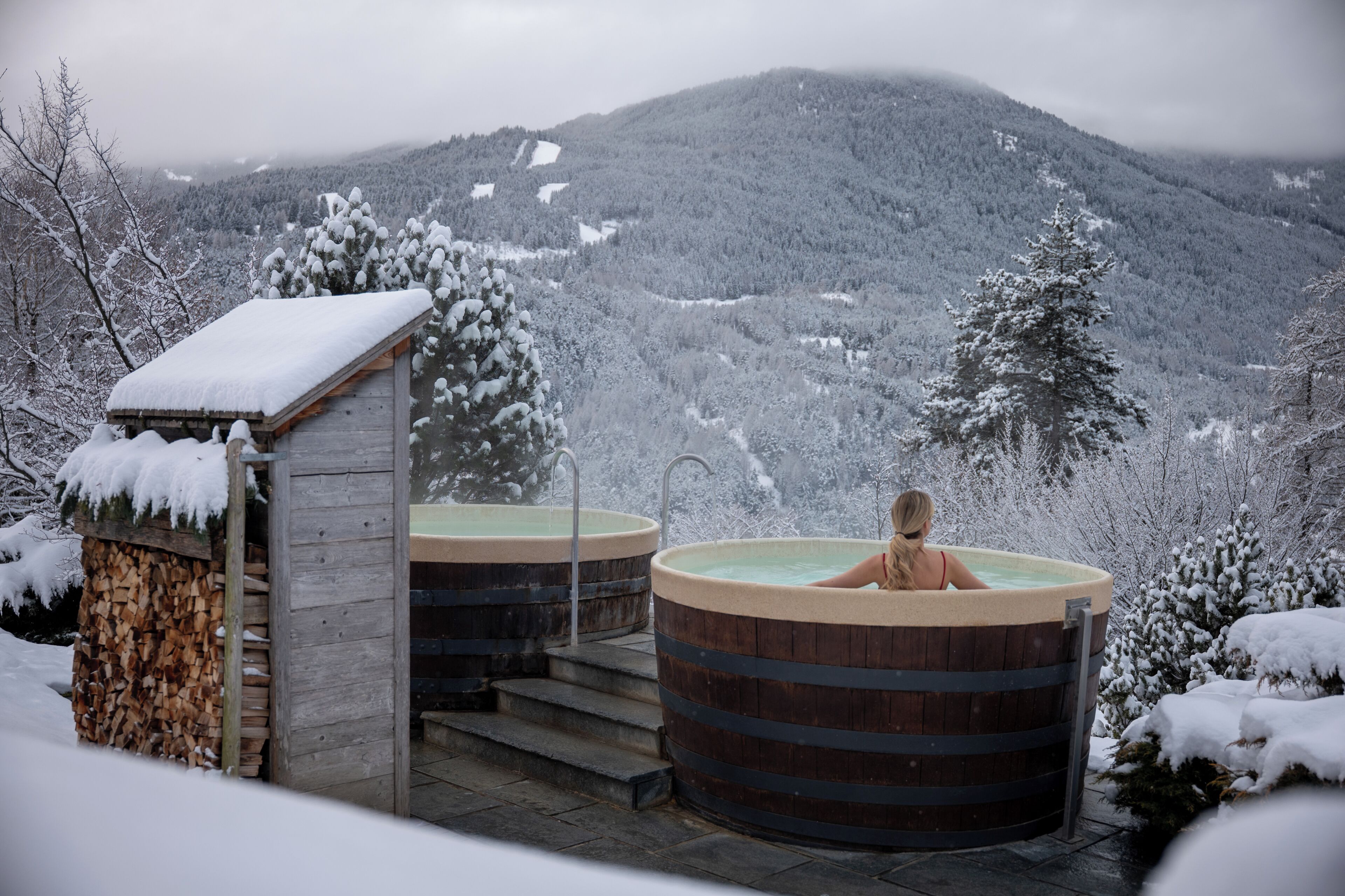 Una cabaña de madera con sauna y bañera de hidromasaje, rodeada de árboles y montañas cubiertas de nieve.