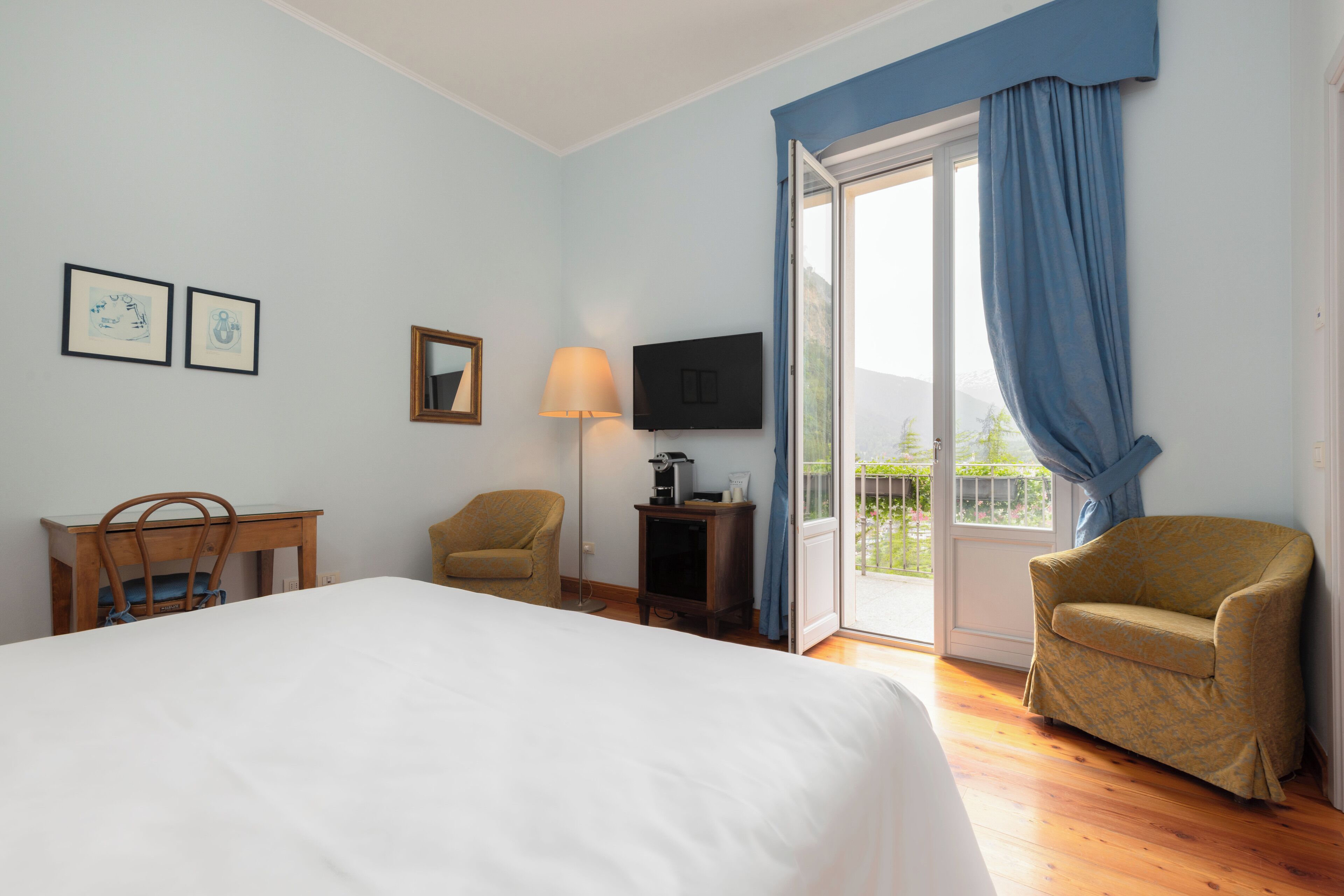 A hotel room with a bed, a desk, a chair, a TV, and a balcony with curtains.