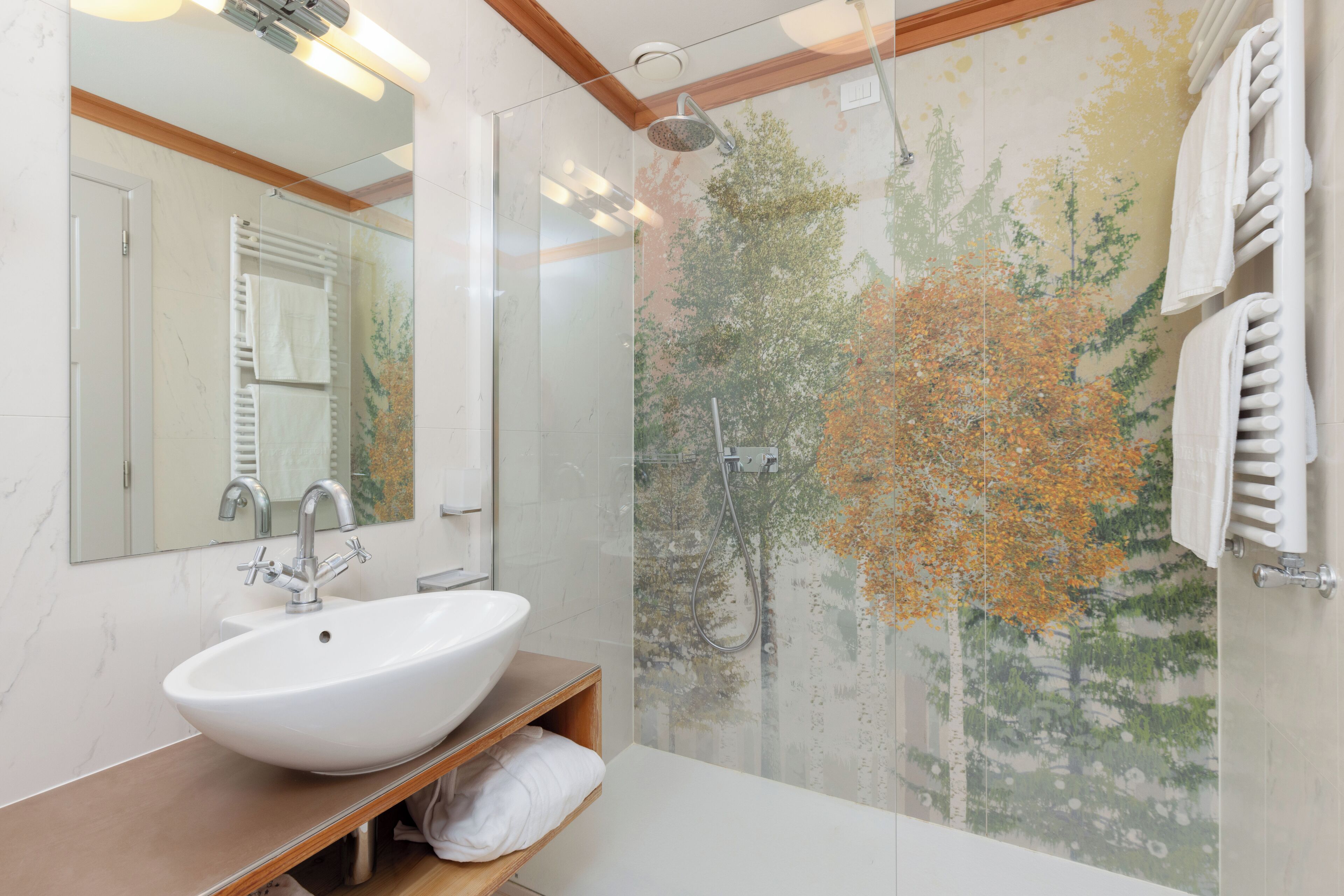 Ein modernes Badezimmer mit einem großen Spiegel, einem weißen Waschbecken, einer Dusche mit Glastür und einer Wand mit einem Baum-Wandbild.