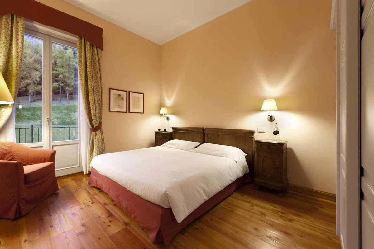 Ein Schlafzimmer mit einem großen Bett, zwei Nachttischen mit Lampen, Holzboden und einem Fenster mit Vorhängen.