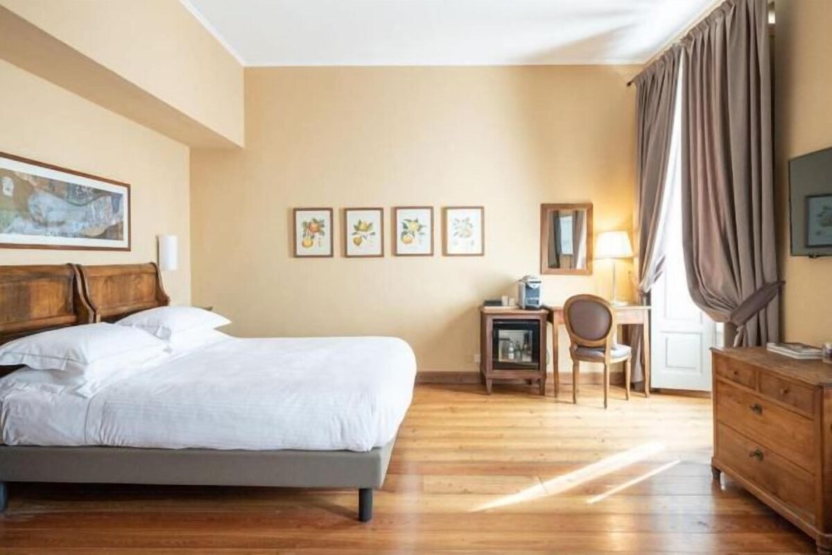 A hotel room with a bed, a desk, a chair, a dresser, and a window with curtains.