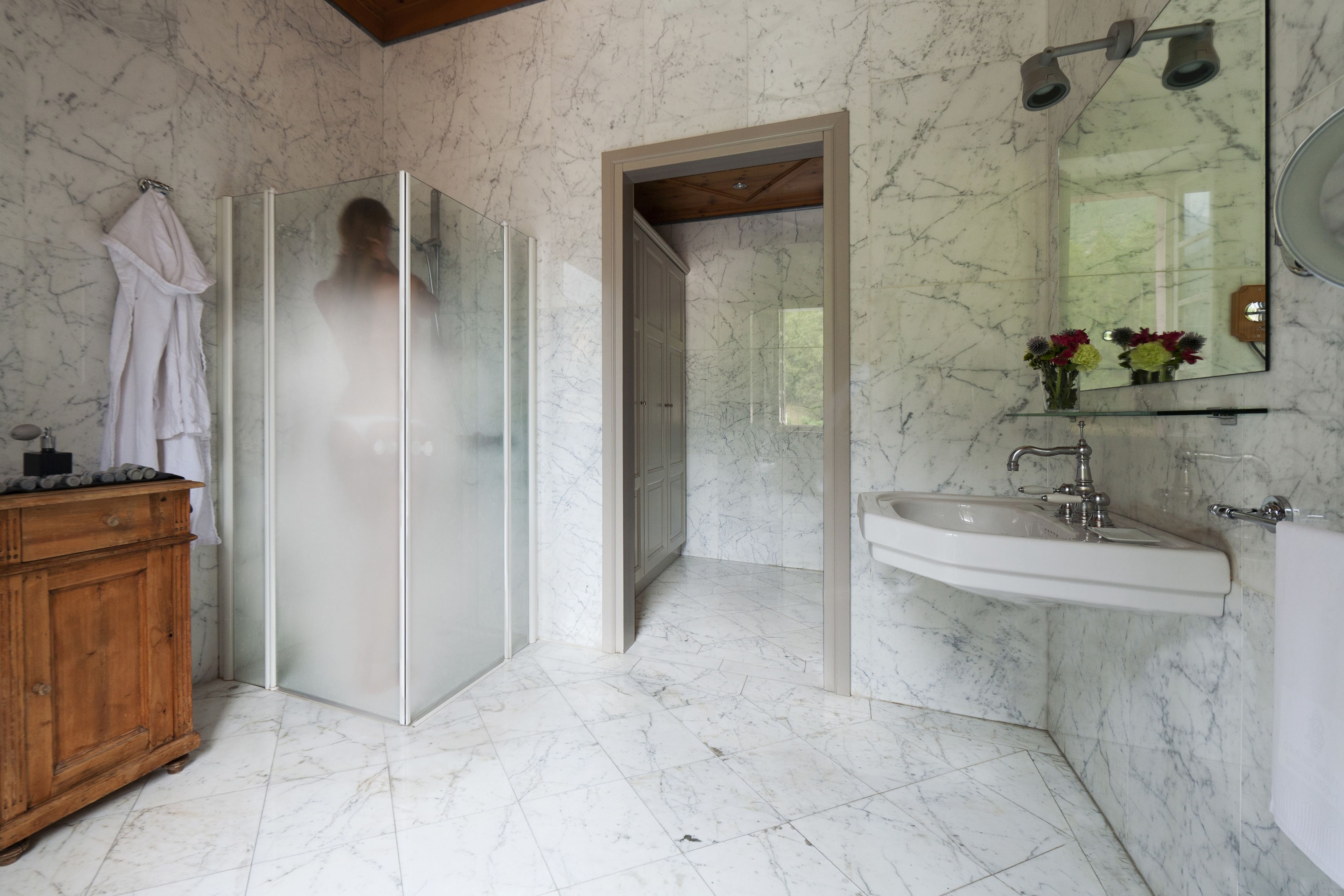 A bathroom with a glass-enclosed shower, a wooden vanity, a marble-tiled floor, a sink, and a mirror with flowers.