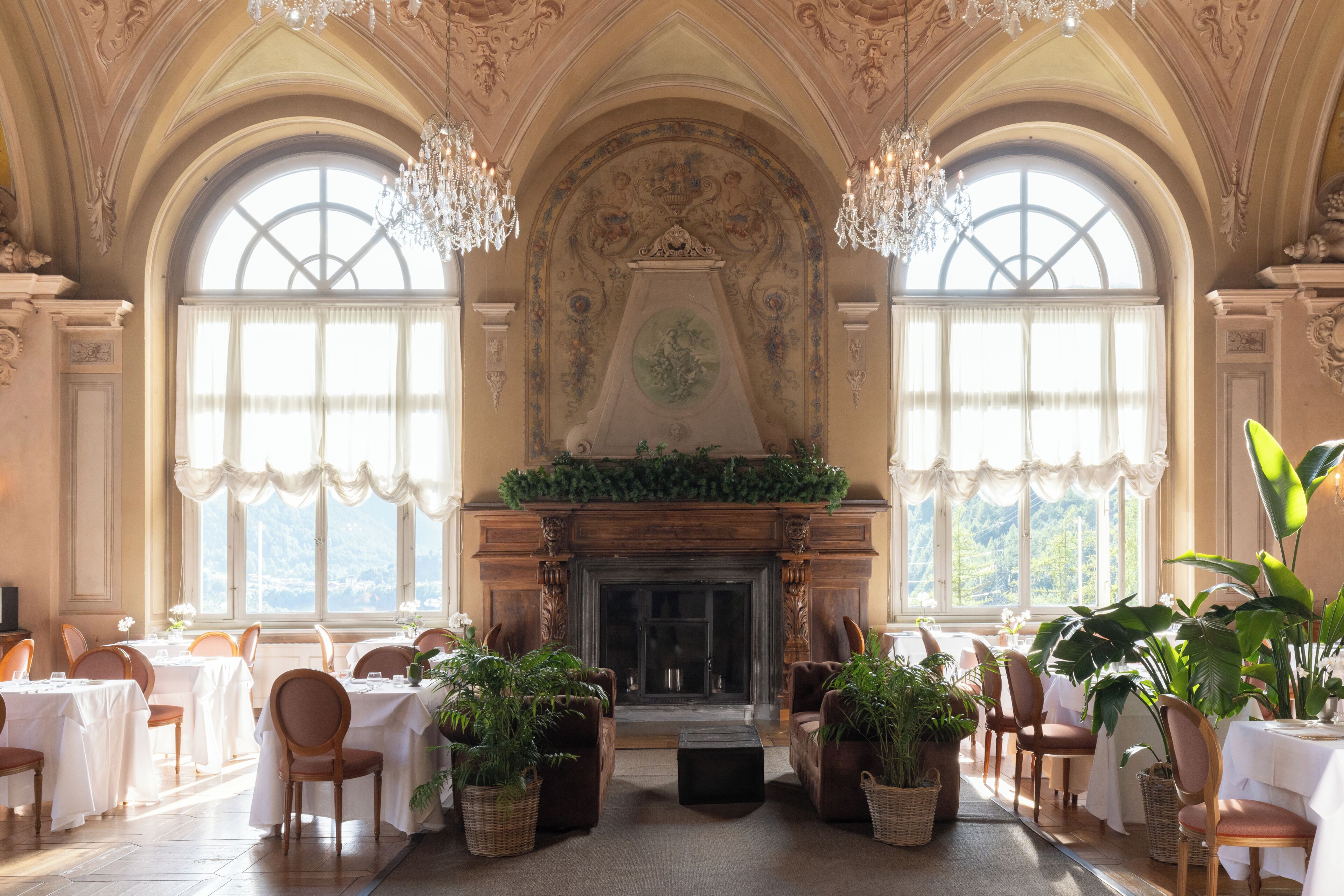 A grand room with a fireplace, large windows, chandeliers, and tables set for dining.