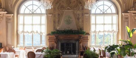 A grand room with a fireplace, large windows, chandeliers, and tables set for dining.
