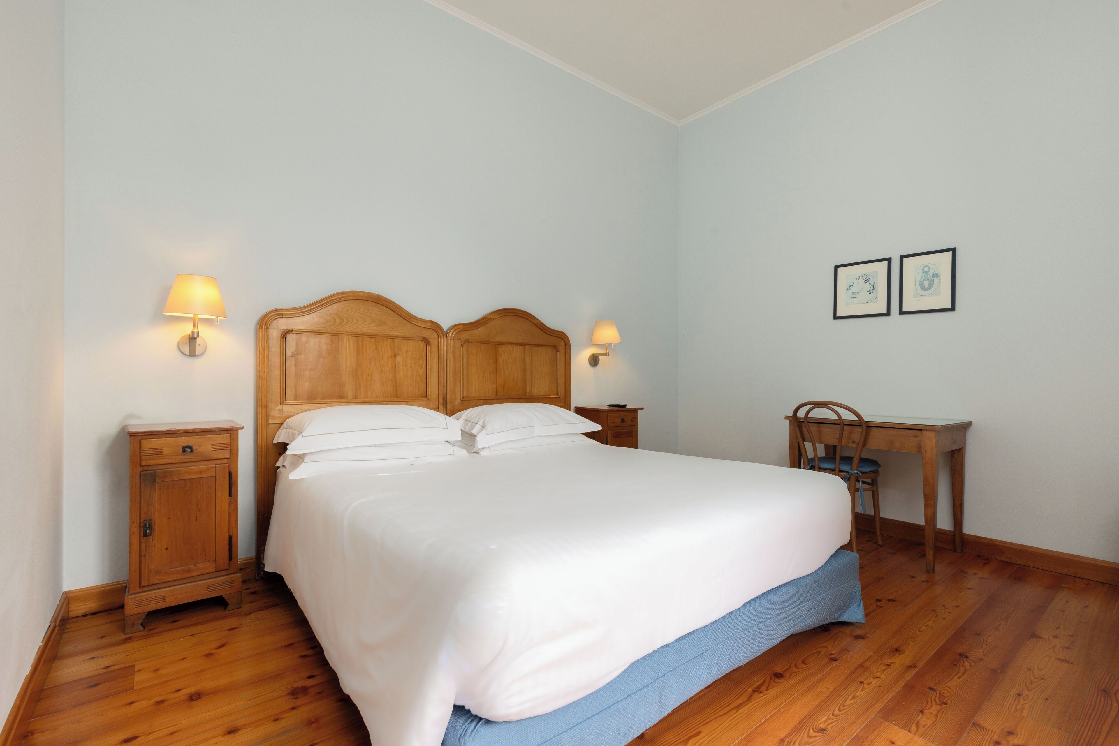 A neatly made bed with a wooden headboard, two bedside tables with lamps, a desk with a chair, and two framed pictures on the wall.