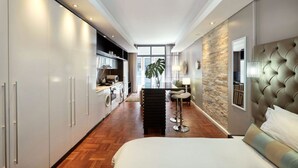 A modern hotel room with a large bed, a seating area with a high backrest, a kitchen area with a washing machine, and a view of the city through large windows. - Apartments at Mandela Rhodes Place (Cape Town)