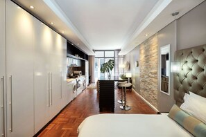Une chambre d’hôtel moderne dotée d’un grand lit, d’un coin salon avec un haut dossier, d’un coin cuisine avec un lave-linge et offrant une vue sur la ville grâce à de grandes fenêtres.