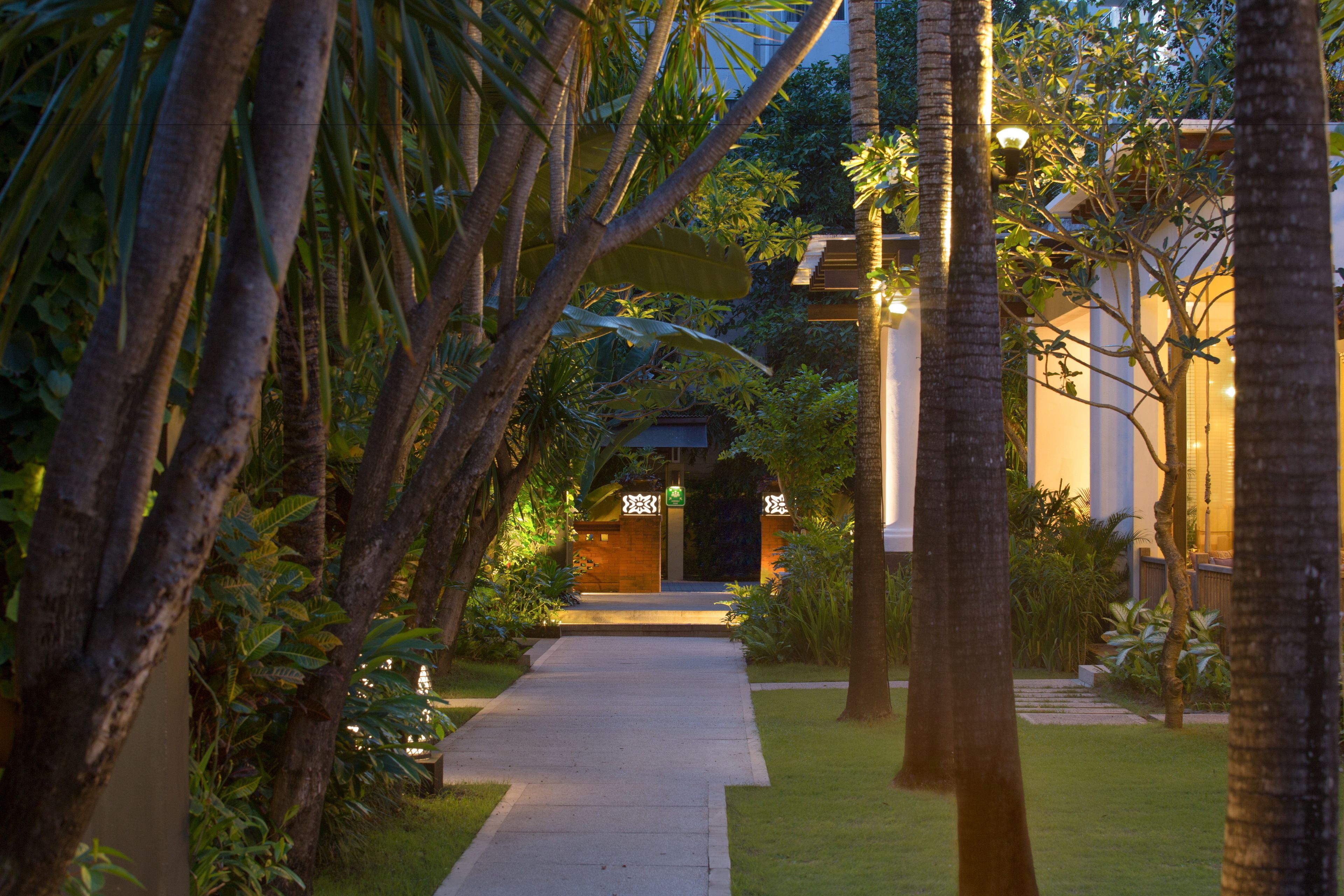 A pathway leading to a building with a gated entrance, surrounded by lush greenery and palm trees.