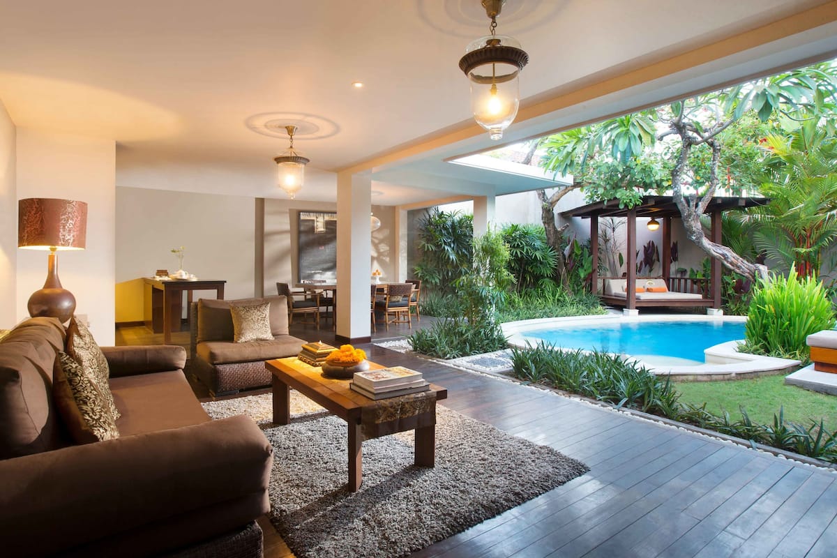 A modern living room with a sofa, coffee table, and lamp. There is a pool area with a slide and lush greenery.
