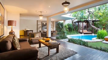A modern living room with a sofa, coffee table, and lamp. There is a pool area with a slide and lush greenery.