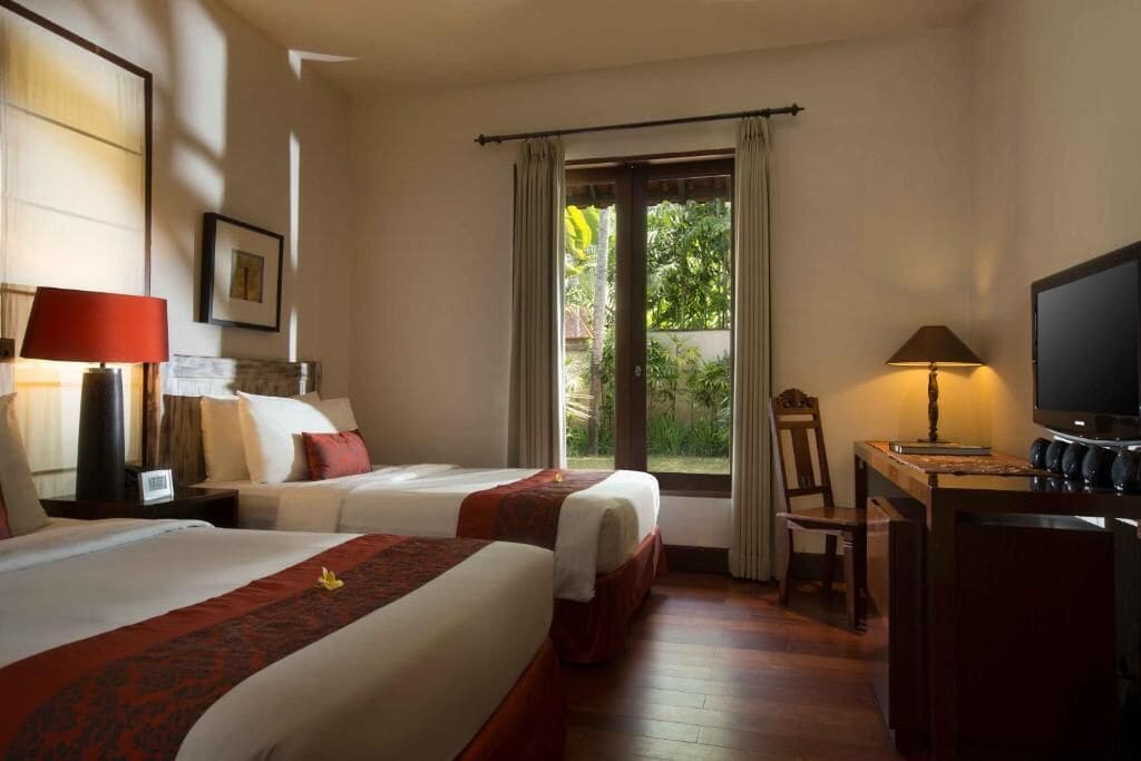 A hotel room with two beds, a desk with a television, and a view of greenery outside.