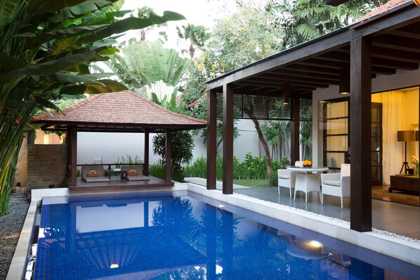 Une piscine avec une terrasse carrelée, un espace détente couvert avec des chaises blanches, et une végétation luxuriante qui entoure l’ensemble.