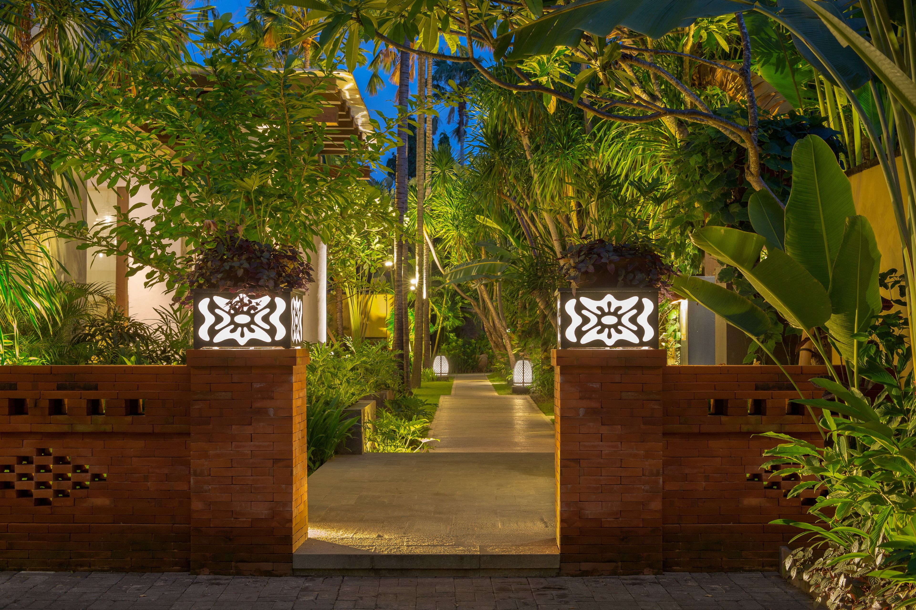 A pathway leading through a lush garden with a brick wall on one side and a white decorative gate on the other.