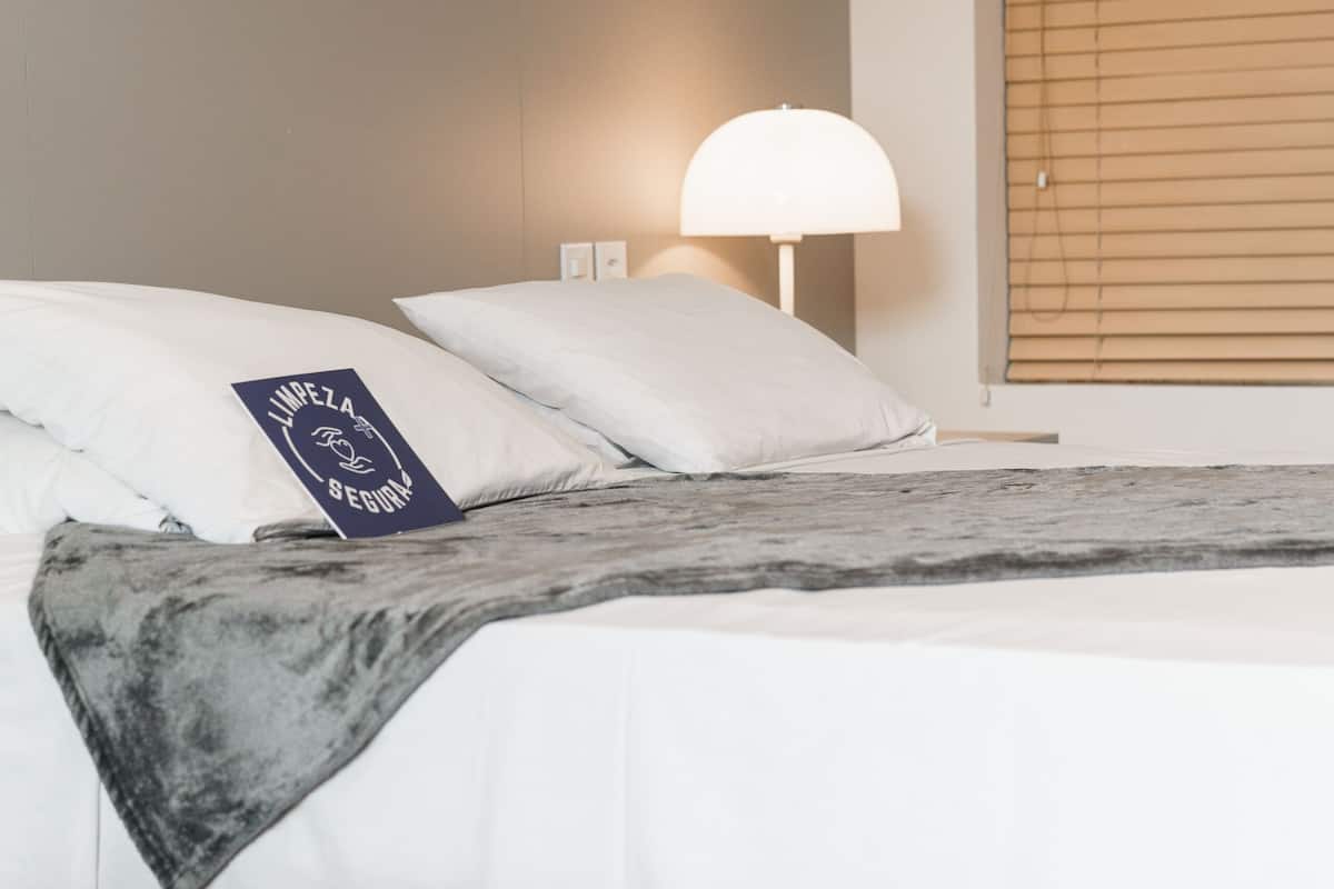 A neatly made bed with white bedding, a dark throw blanket, and a pillow with text. There is a wall-mounted lamp and a window with blinds.