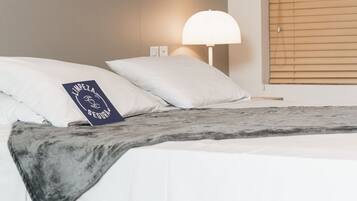 Een net opgemaakt bed met witte lakens, een donkere plaid en een kussen met tekst. Er is een wandlamp en een raam met zonwering.