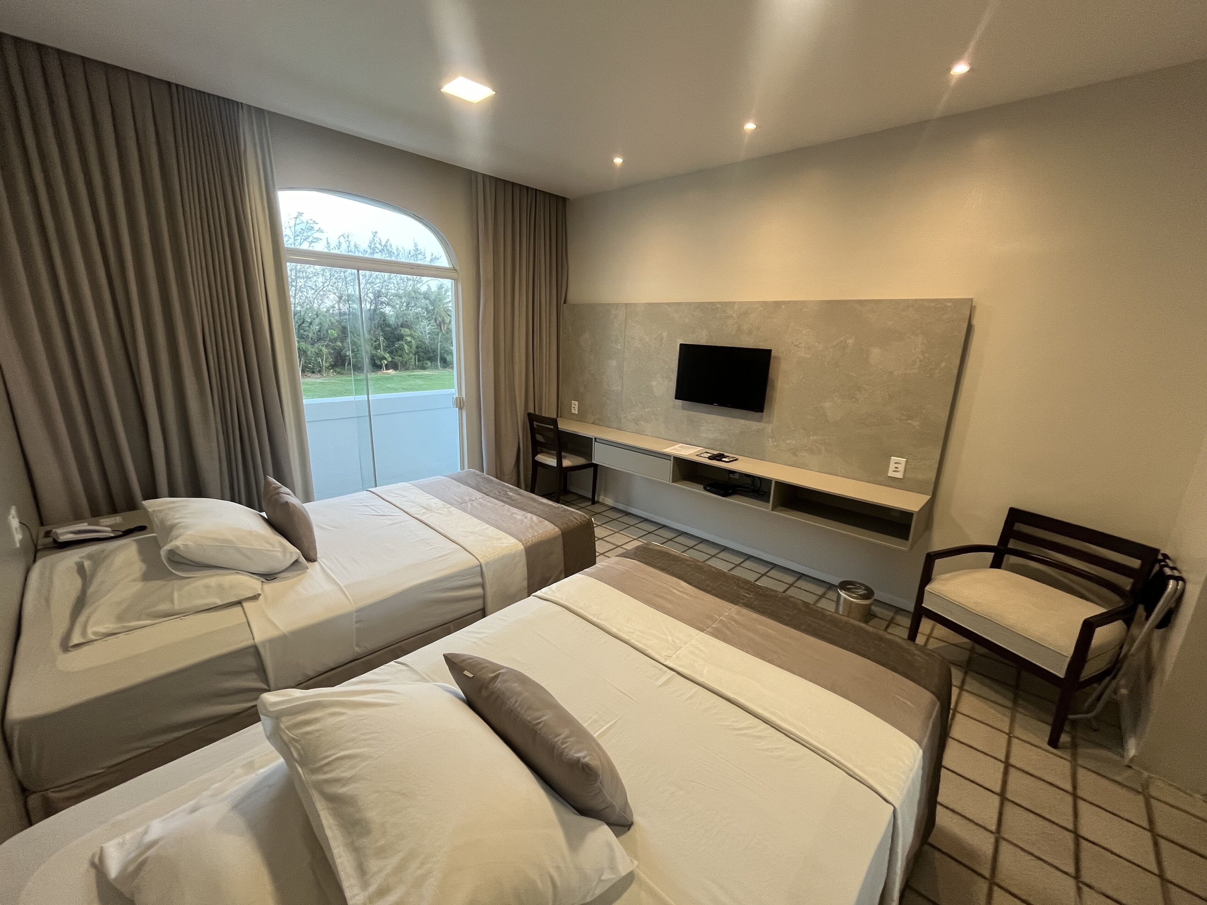 Une chambre d’hôtel avec deux lits, un téléviseur fixé au mur, un bureau avec une chaise et une fenêtre avec des rideaux.