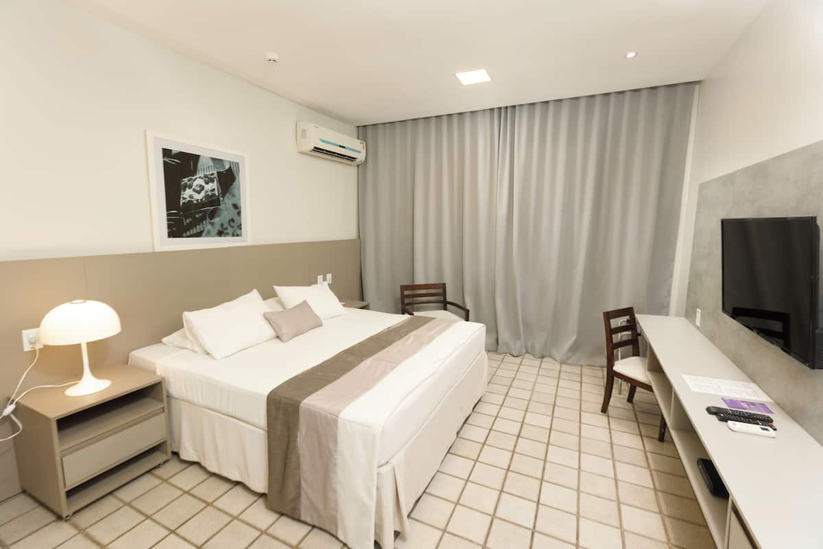 A hotel room with a bed, bedside table, lamp, desk, chair, TV, and curtains.