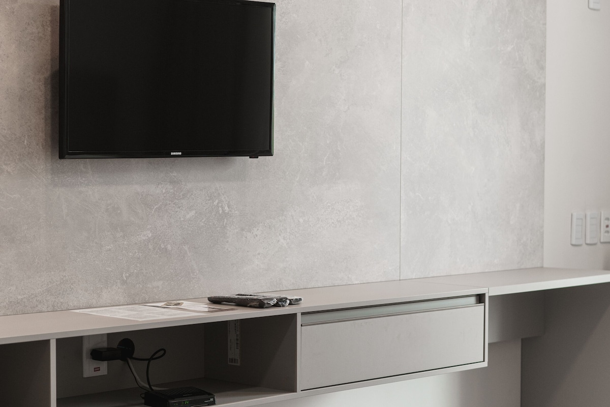 Una habitación moderna con un televisor de pantalla plana sujeto a una pared con textura, una mesita de noche blanca con cajón y una cama con cubrecama oscuro.
