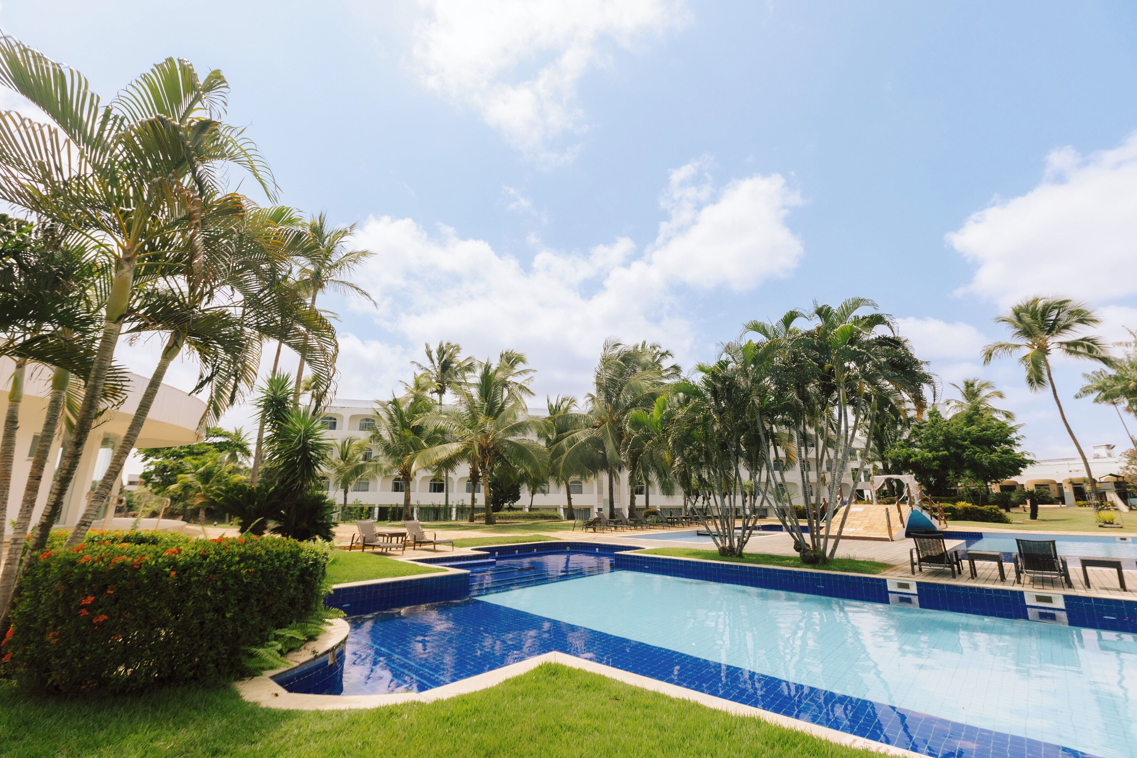 A swimming pool with a blue tiled edge, surrounded by palm trees and greenery, with lounge chairs and a building in the background.