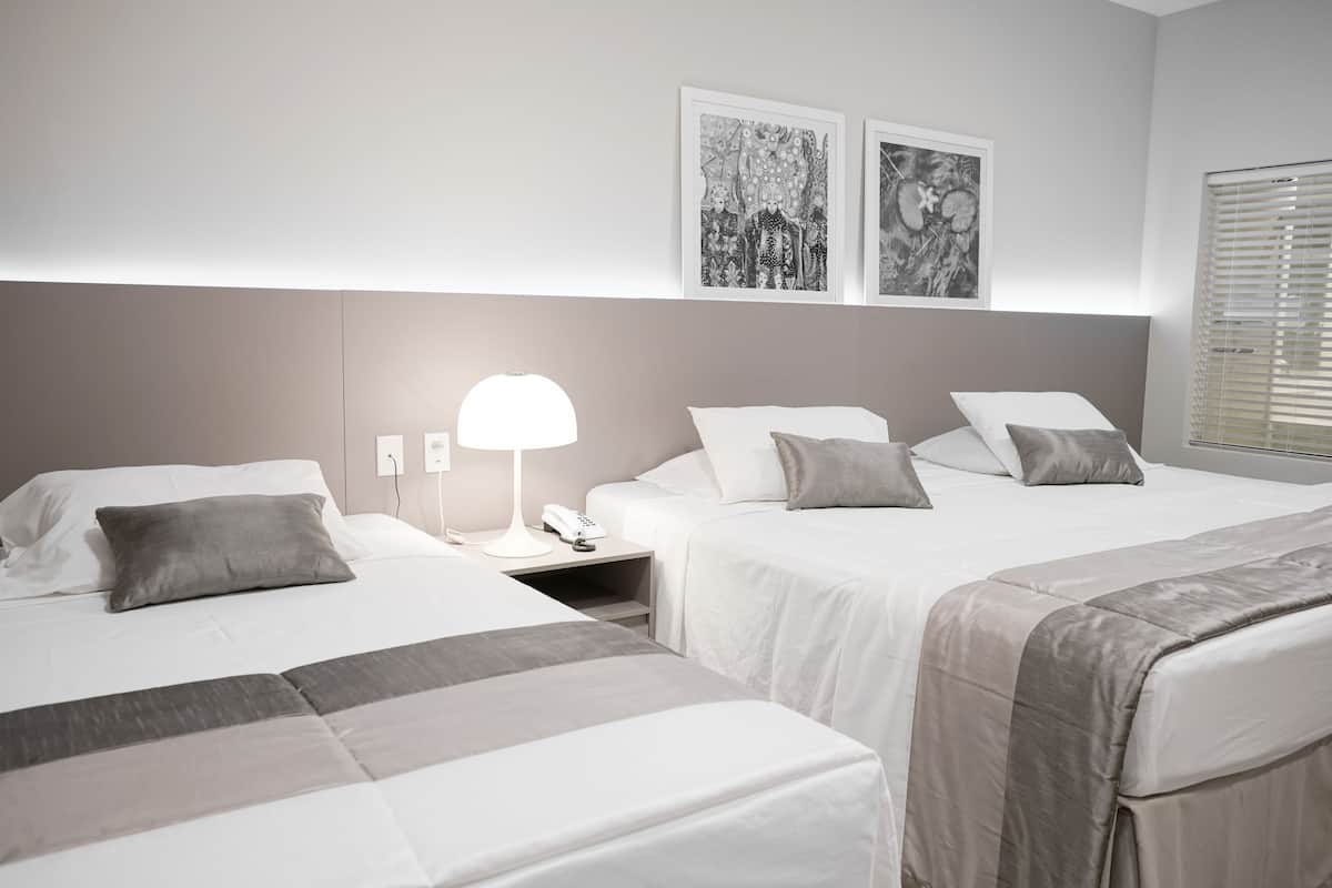A hotel room with three beds, a bedside table with a lamp, and two framed artworks on the wall.
