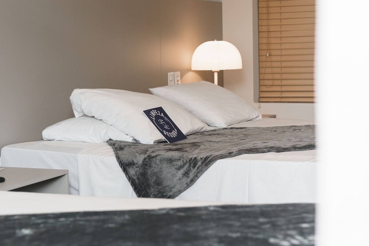 A neatly made bed with white bedding and a black and white throw blanket. There is a bedside table with a lamp and a small rectangular object on it.