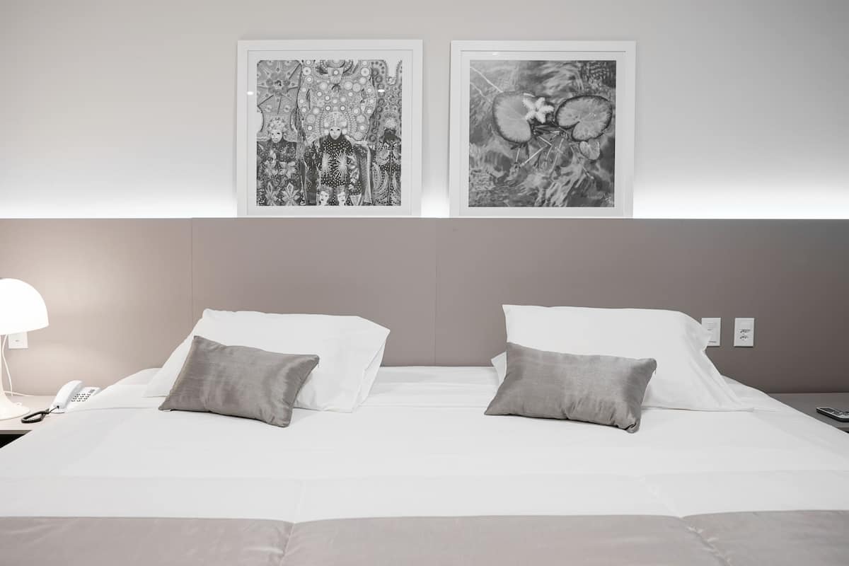 A hotel room with a large bed, two pillows, a bedside lamp, a phone, and two framed artworks on the wall.