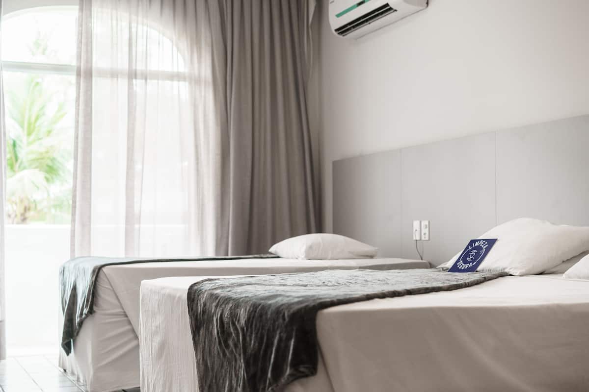 A hotel room with two beds, a large window with sheer curtains, and an air conditioning unit on the wall.