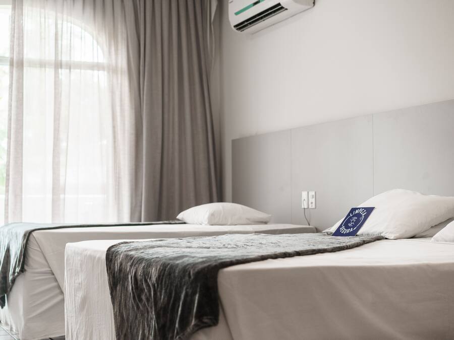 A hotel room with two beds, a large window with sheer curtains, and an air conditioning unit on the wall.