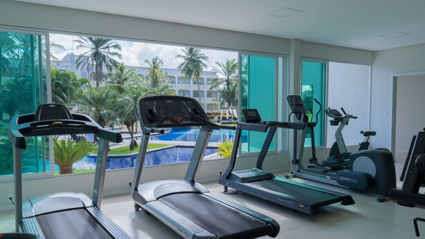 Une salle de sport équipée de tapis de course, d’un vélo elliptique et d’un rameur, avec vue sur une piscine et des palmiers.